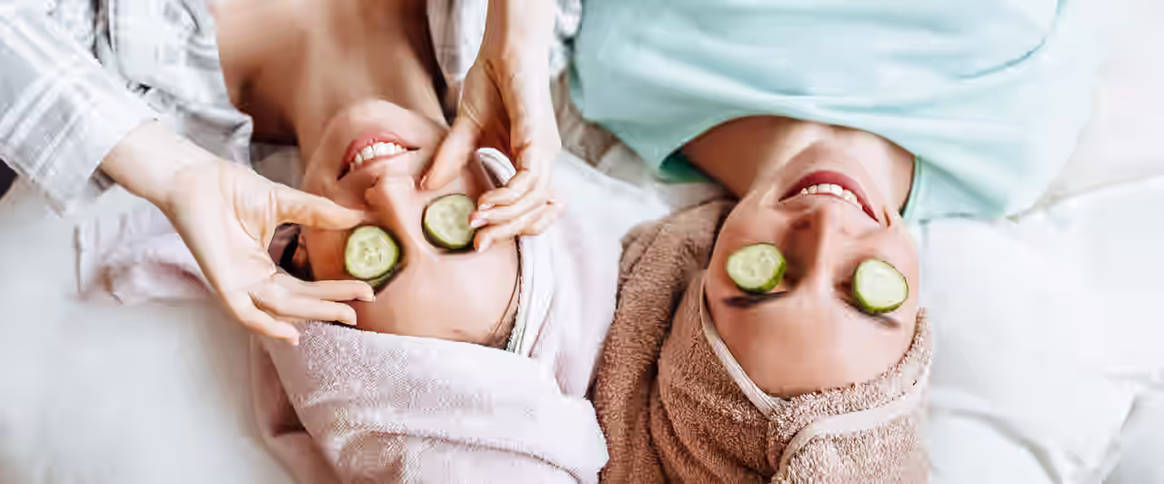 2 Frauen auf einem Bett mit Handtüchern auf dem Kopf und Gurkenscheiben auf den Augen