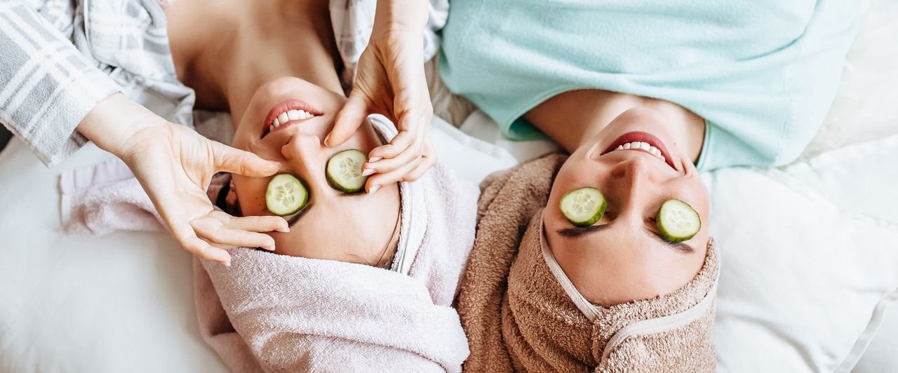 2 Frauen auf einem Bett mit Handtüchern auf dem Kopf und Gurkenscheiben auf den Augen
