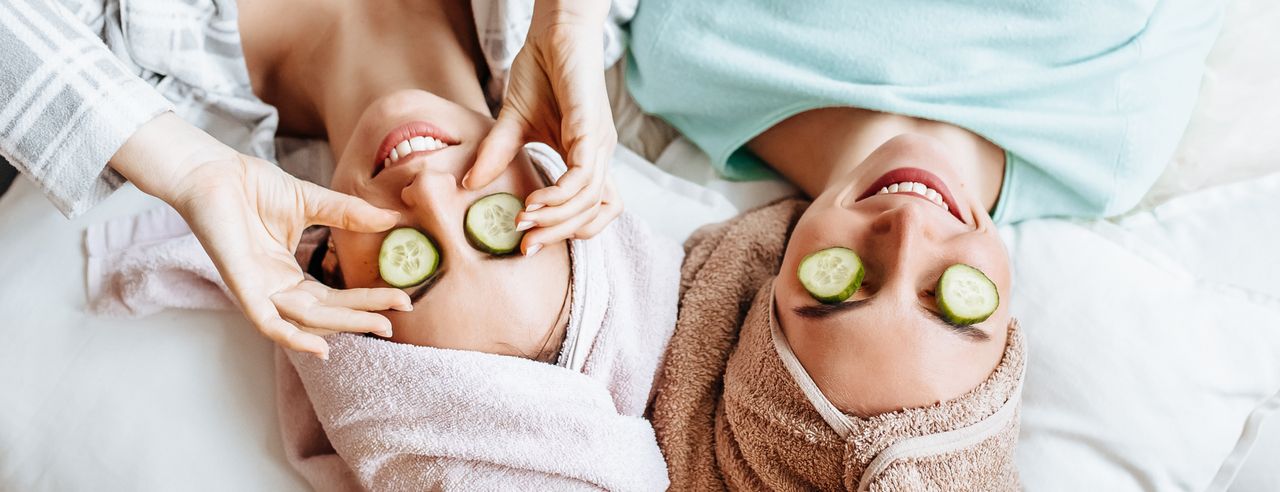 2 Frauen auf einem Bett mit Handtüchern auf dem Kopf und Gurkenscheiben auf den Augen