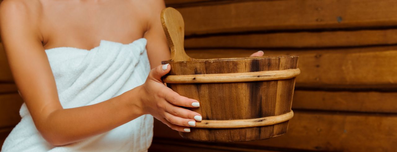 Eine Frau sitzt mit einem weißen Handtuch bekleidet in der Sauna und hält einen Holzeimer in den Händen.