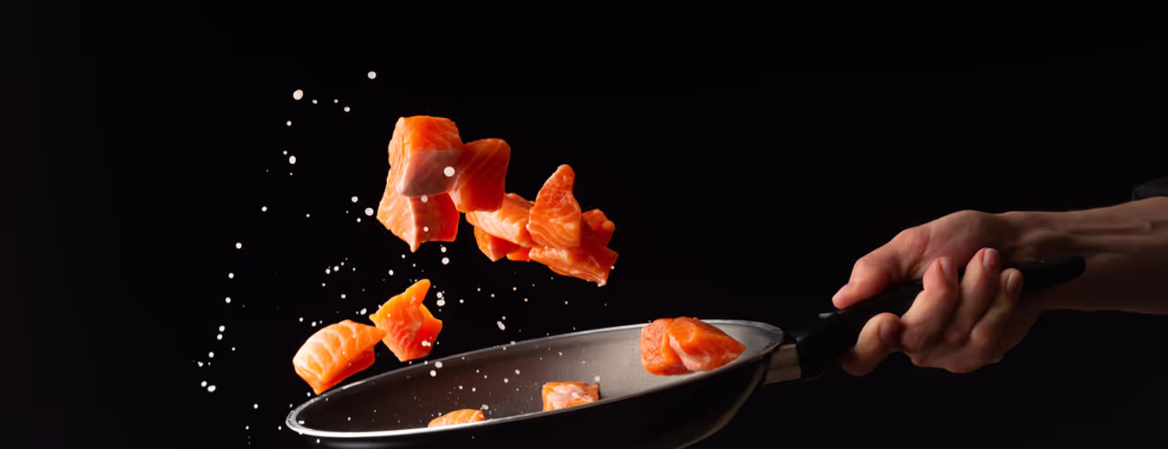 Eine Hand schwenkt Lachsstücke in einer Pfanne, vor schwarzem Hintergrund