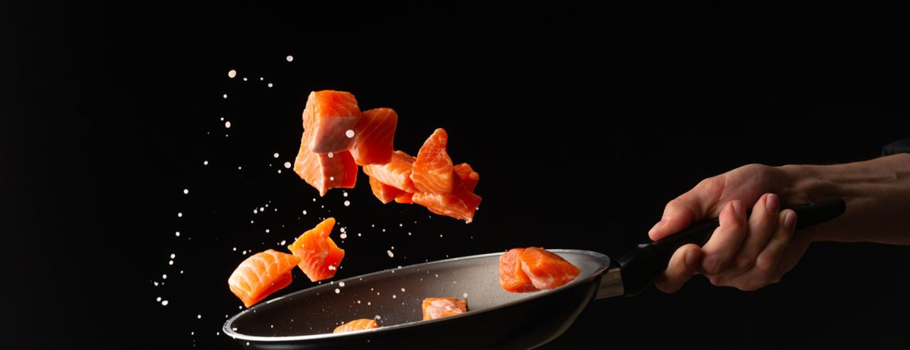 Eine Hand schwenkt Lachsstücke in einer Pfanne, vor schwarzem Hintergrund