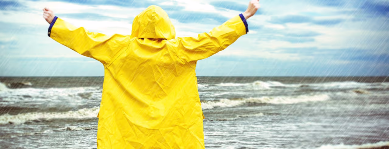 Frau im gelben Parker steht mit ausgebreiteten Armen im Regen am Meer