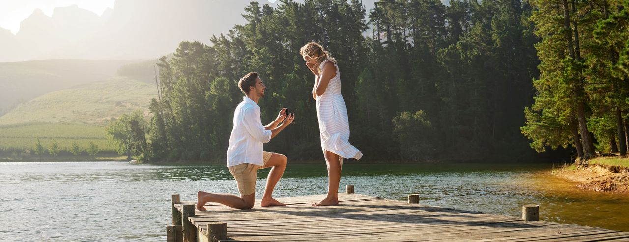 Ein Mann präsentiert einer Frau auf Knien einen Verlobungsring auf dem Steg eines Sees