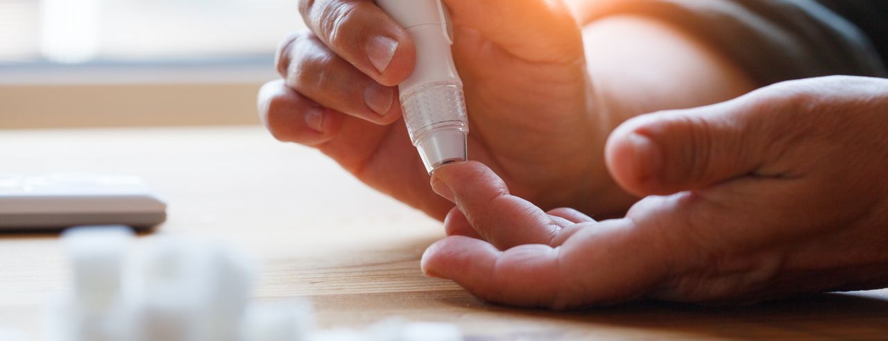 Eine Hand hält einen Blutzuckermessgerät-Pen, während ein Finger der anderen Hand vorbereitet wird, um eine Blutprobe zu entnehmen. Im Vordergrund unscharf sind Zuckerwürfel zu sehen.