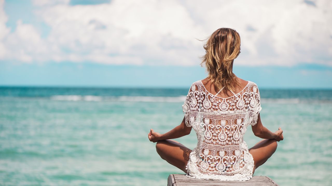 Eine Frau in weißer Yoga Kleidung sitzt im Lotussitz an der Küste Kroatiens und schaut auf das Meer hinaus