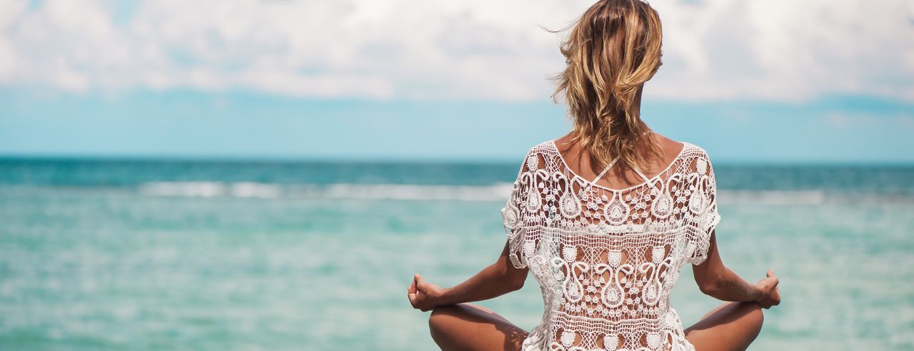 Eine Frau in weißer Yoga Kleidung sitzt im Lotussitz an der Küste Kroatiens und schaut auf das Meer hinaus