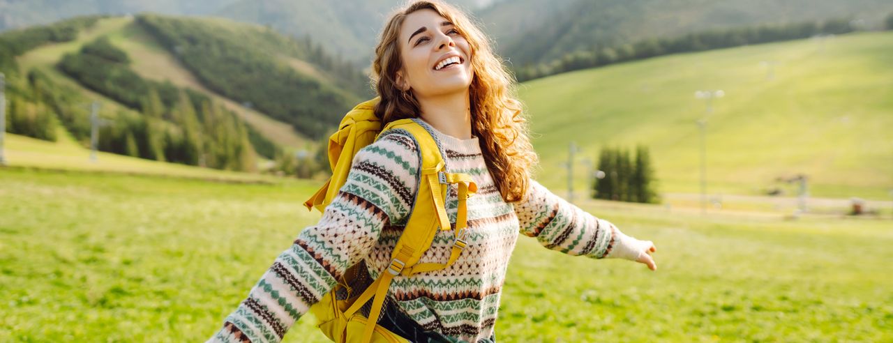 Eine Frau mit gelbem Rucksack steht auf einer grünen Wiese und streckt freudig die Arme zu den Seiten aus.