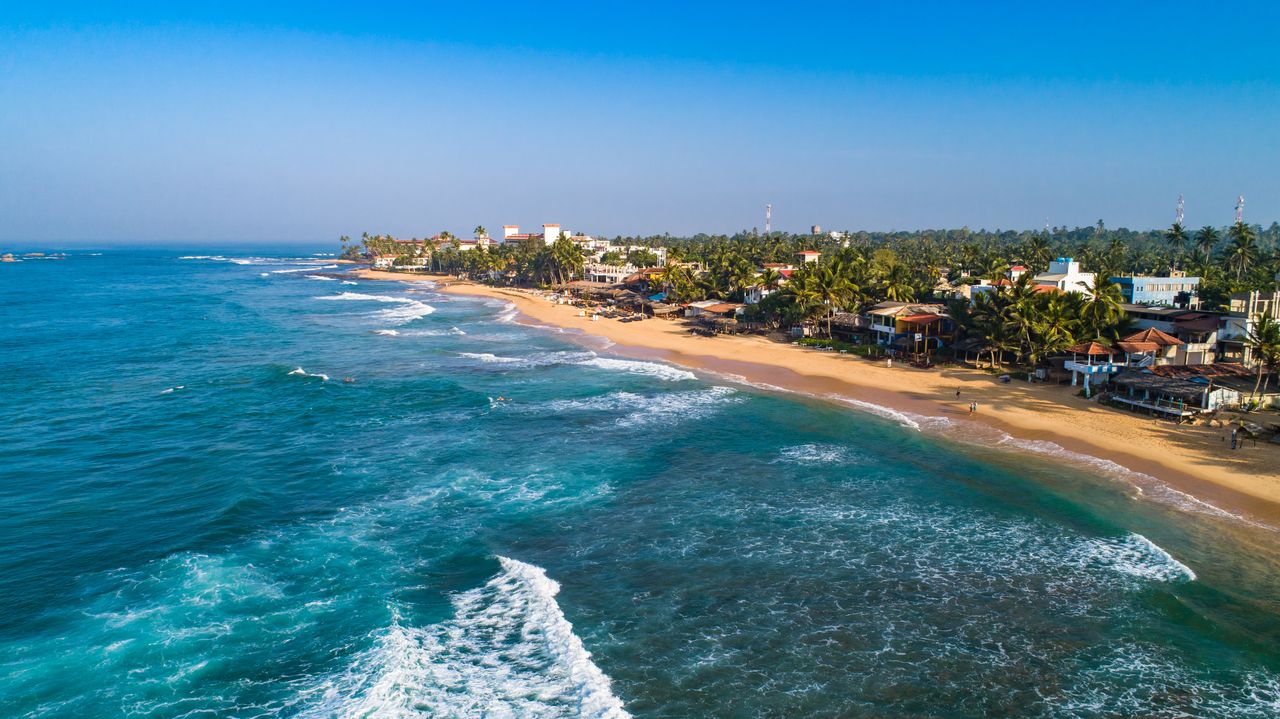 Luftaufnahme von blauem Meer mit weißen Wellen vor dem goldenen Sandstrand von Hikkaduwa mit Gebäuden zwischen Palmen