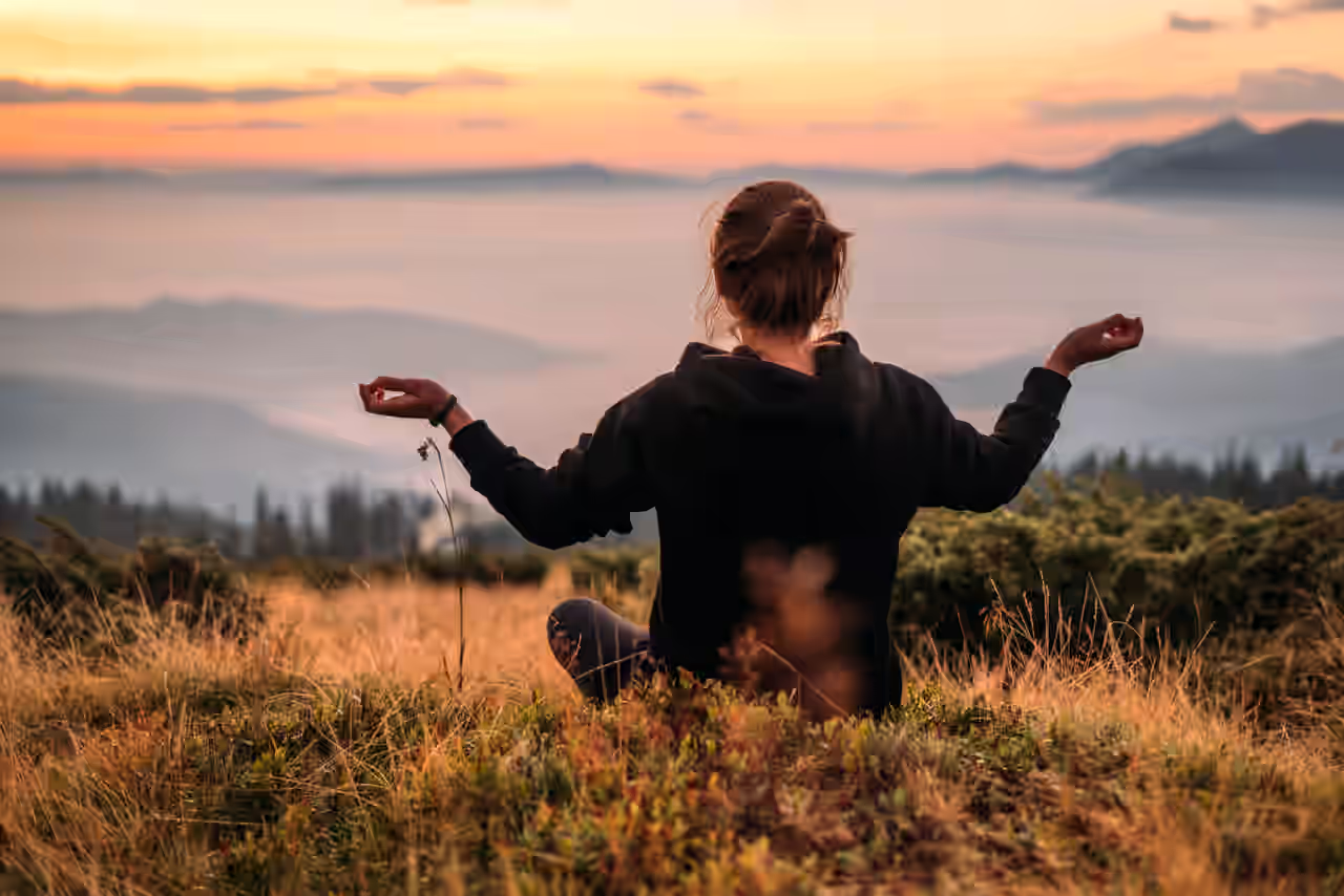 Eine Frau sitzt in Meditationshaltung auf einer Weise und blickt über die Landschaft
