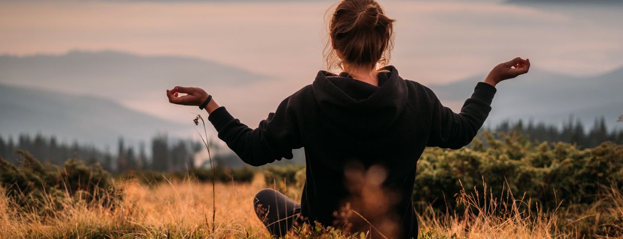 Eine Frau sitzt in Meditationshaltung auf einer Weise und blickt über die Landschaft