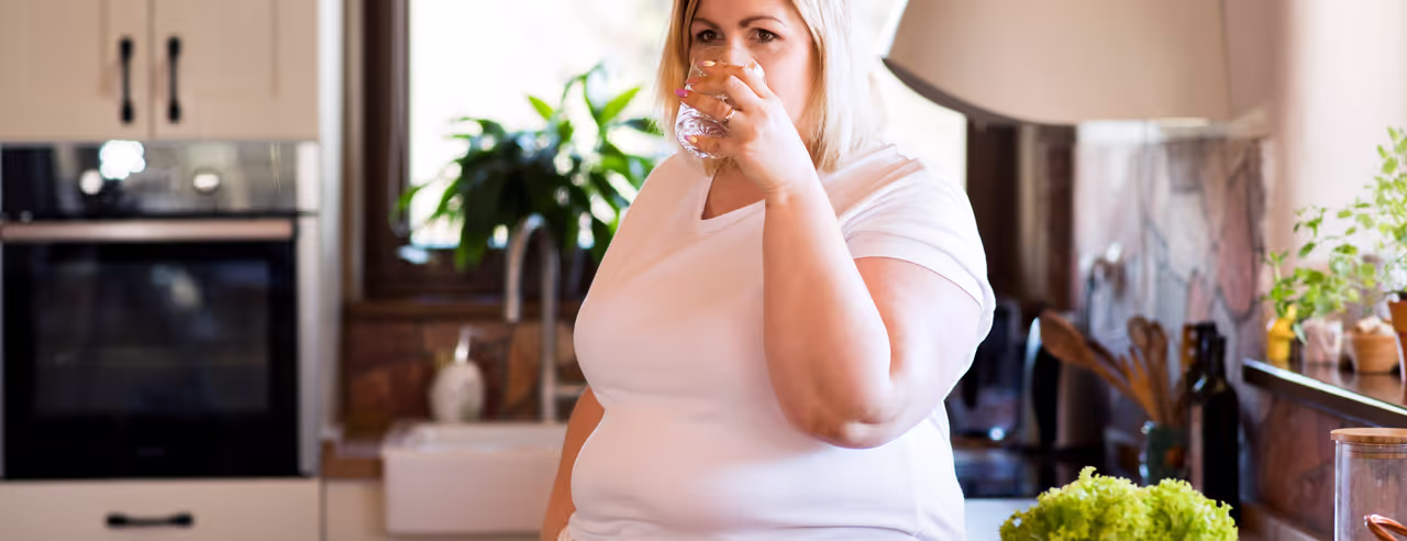 Übergewichtige Frau trinkt in der Küche ein Glas Leitungswasser neben eine Tresen mit Gemüse für Salat