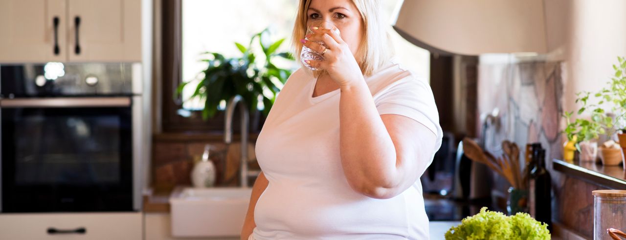 Übergewichtige Frau trinkt in der Küche ein Glas Leitungswasser neben eine Tresen mit Gemüse für Salat