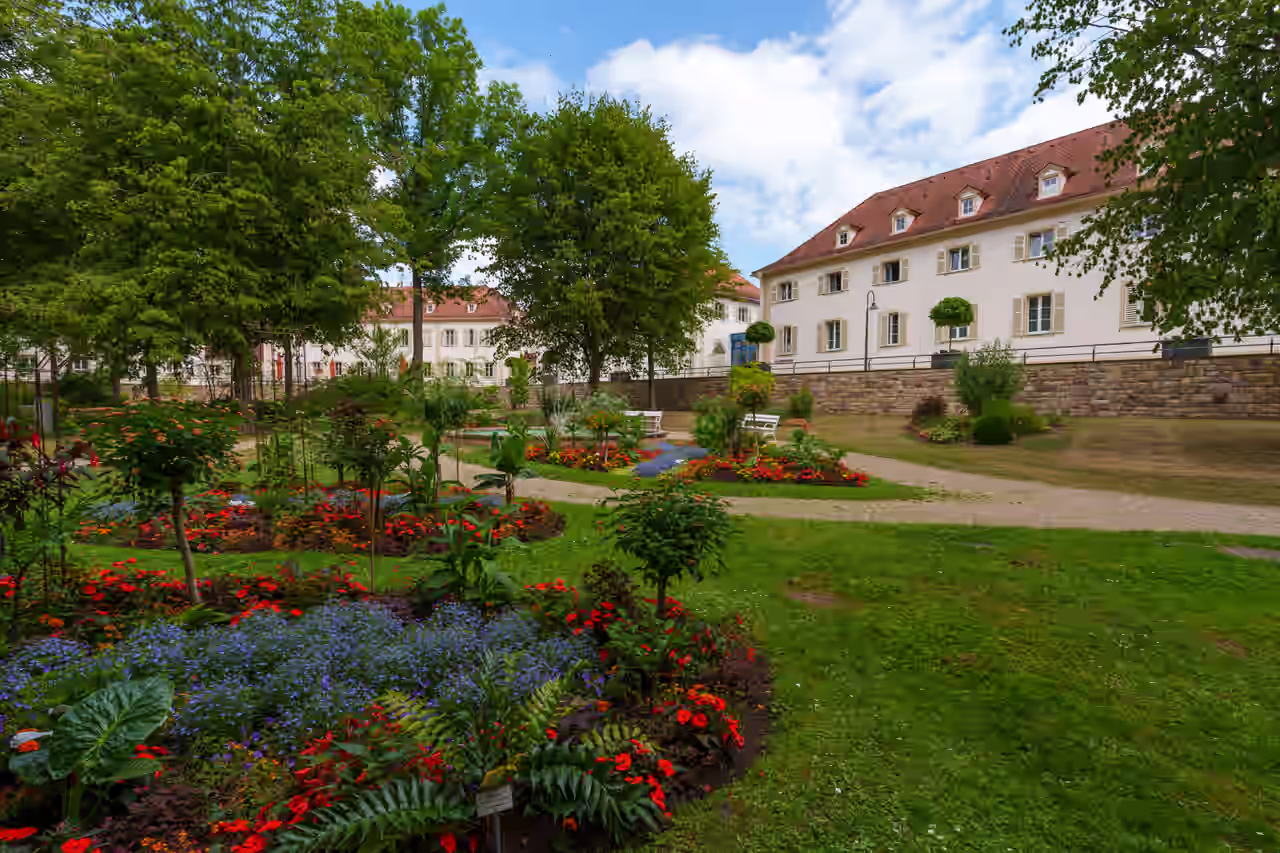 Farbenfrohe Beete auf grünem Rasen zwischen Bäumen und Kieswegen im Kurpark Bad Bocklet vor dreistöckigen hellen Häusern hinter einer Mauer mit Geländer