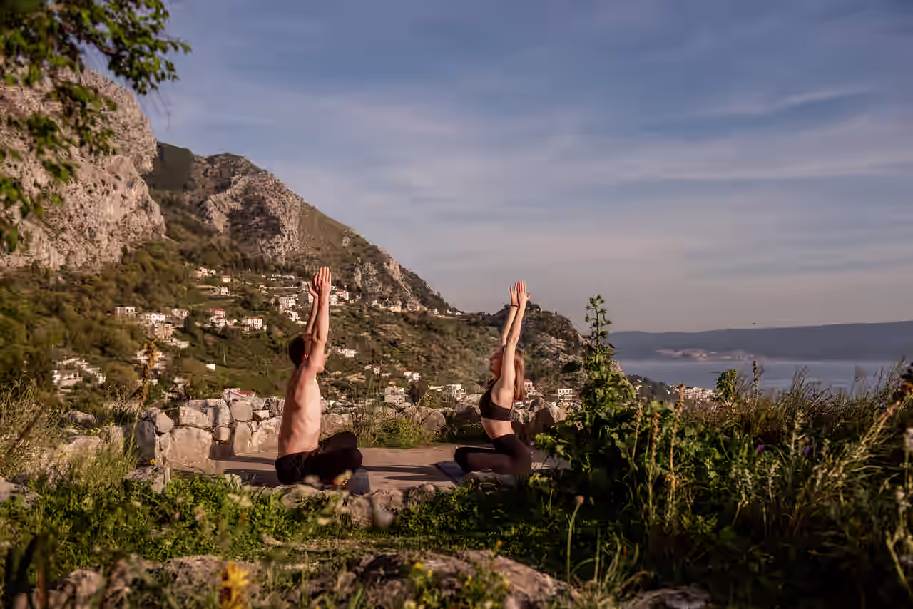Mann und Frau in Yoga Pose in grüner, bergiger Landschaft in Küstennähe