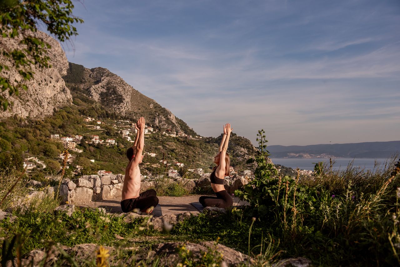 Mann und Frau in Yoga Pose in grüner, bergiger Landschaft in Küstennähe