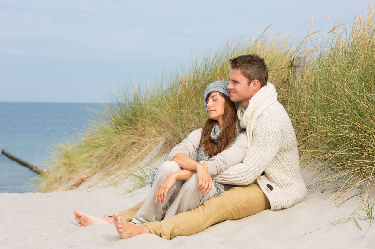 Ein Paar sitzt in den Dünen an der Nordsee. Sie wird von ihm von hinten umarmt und beide schauen während ihrer Flitterwochen auf das Meer.