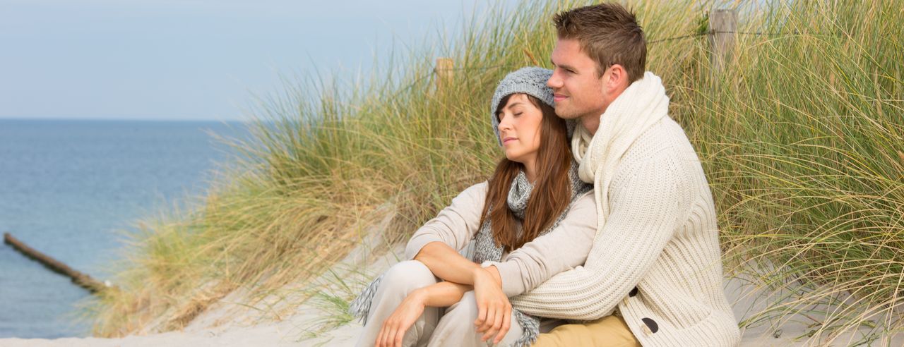 Ein Paar sitzt in den Dünen an der Nordsee. Sie wird von ihm von hinten umarmt und beide schauen während ihrer Flitterwochen auf das Meer.