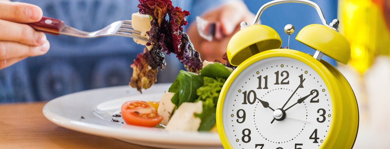 Eine Frauenhand nimmt mit einer Gabel Salat mit Feta von einem Teller neben einem gelben Wecker