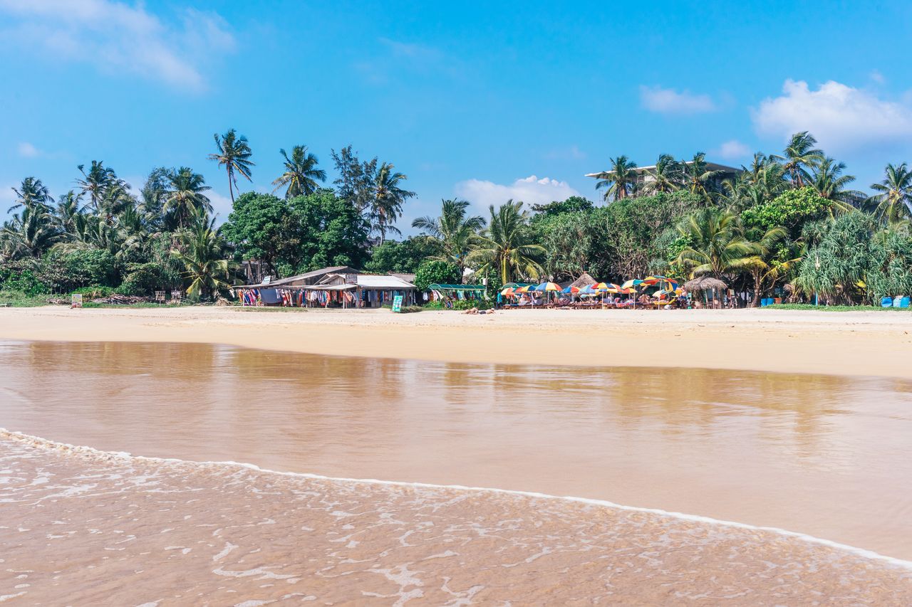 Touristen auf Sonnenliegen unter bunten Schirmen am goldenen Sandstrand von Beruwela neben einem Strandgeschäft mit Bekleidung zwischen Palmen
