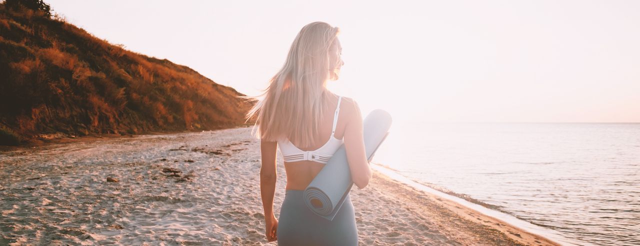 Blonde Frau im Sonnenschein am Strand entlang, mit Yogamatte unter dem Arm