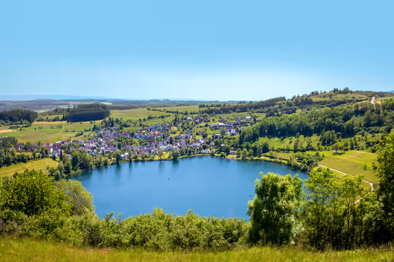 Vulkanischer Maar-See in der Eifel zwischen Bäumen und Wiesen rund um eine Ortschaft