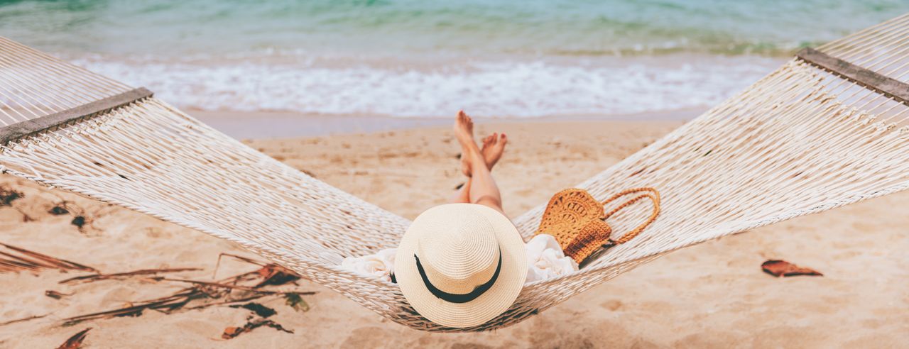 Eine Frau mit weißem Hut liegt am Strand auf einer weißen Hängematte und schaut aufs Meer