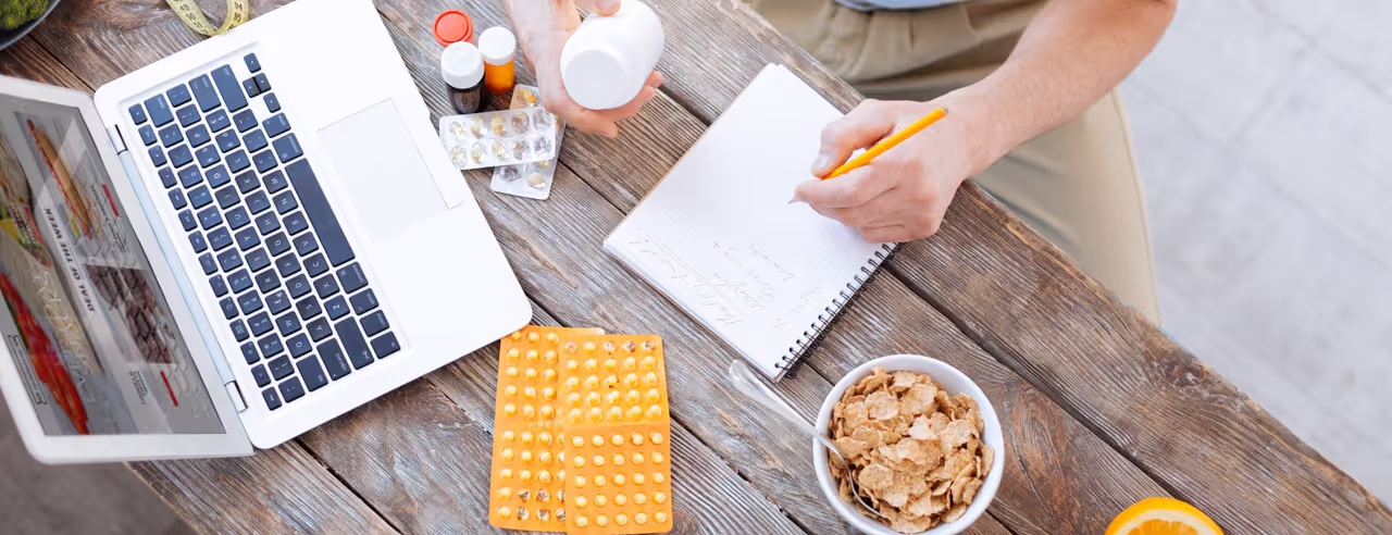 Tisch mit Laptop, Tabletten, Müsli und einem Mann der in ein Notizbuch schreibt.