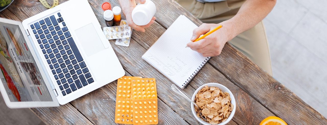 Tisch mit Laptop, Tabletten, Müsli und einem Mann der in ein Notizbuch schreibt.