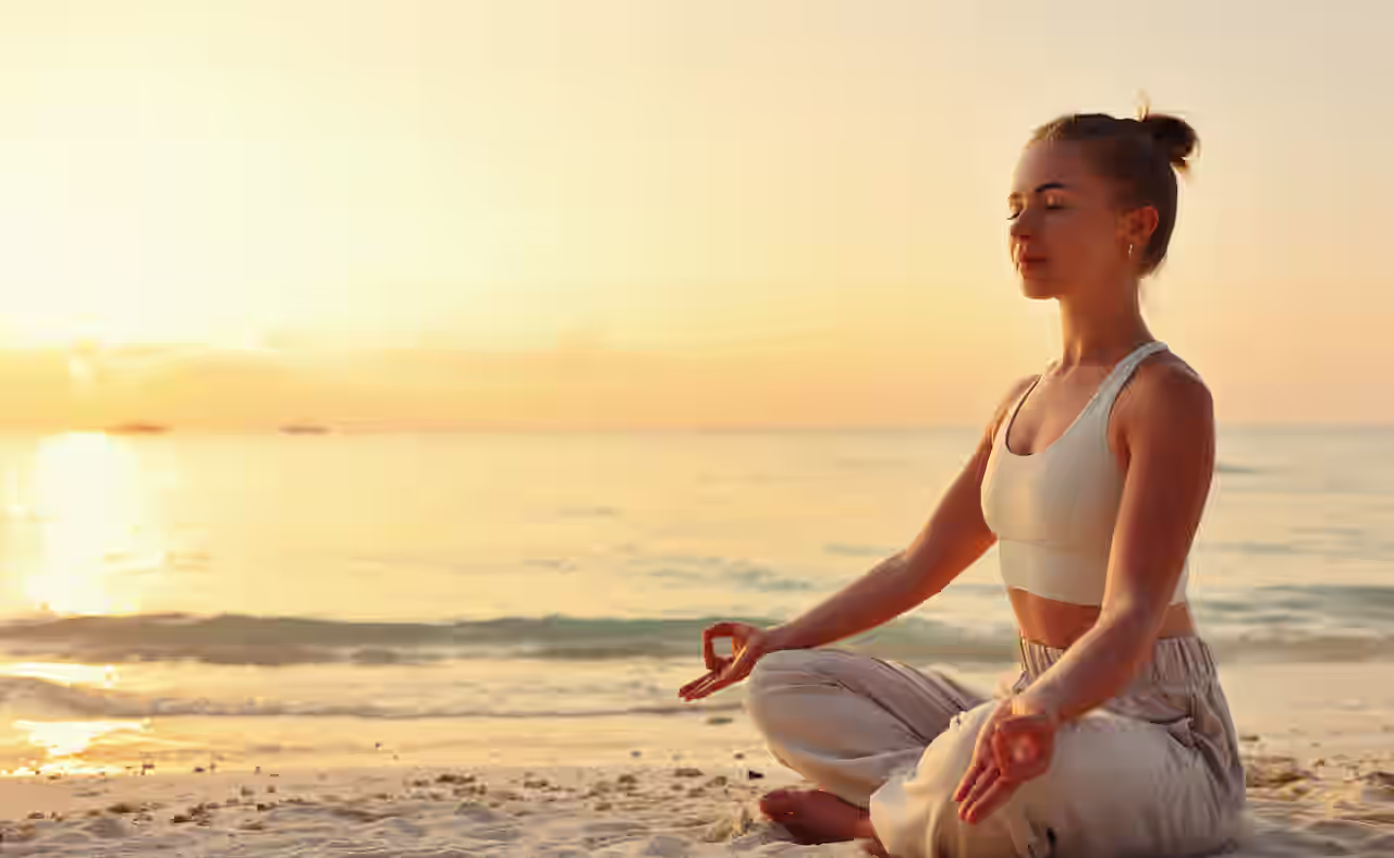 Eine Frau sitzt im Schneidersitz und mit geschlossenen Augen am Strand und meditiert.