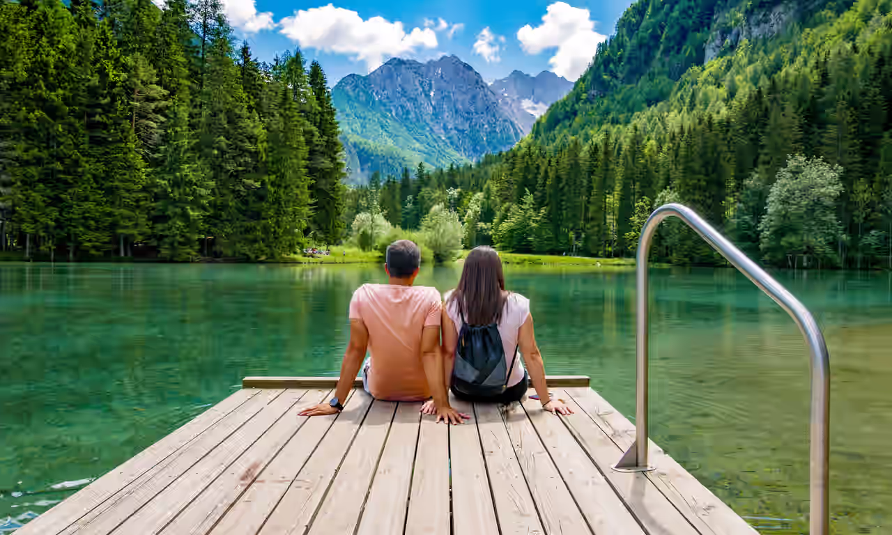 Ein Mann und eine Frau sitzen bei sommerlichen Wetter nebeneinander auf einem Steg und schauen auf einen türkisfarbenen See mit saftig grüner Waldlandschaft im Hintergrund