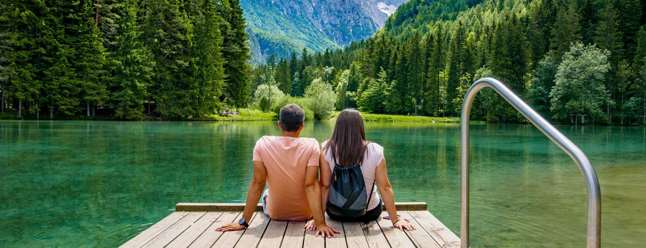 Ein Mann und eine Frau sitzen bei sommerlichen Wetter nebeneinander auf einem Steg und schauen auf einen türkisfarbenen See mit saftig grüner Waldlandschaft im Hintergrund