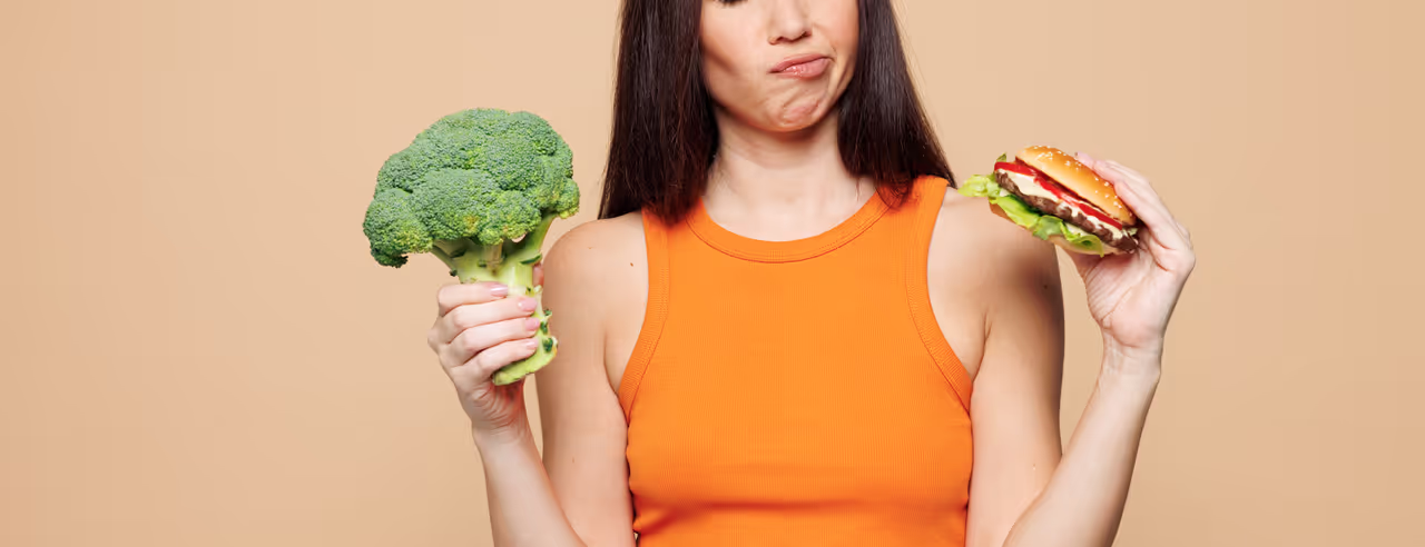 Junge Frau mit rohem Brokkoli in der rechten Hand blickt zweifelnd auf einen Burger in ihrer linken Hand