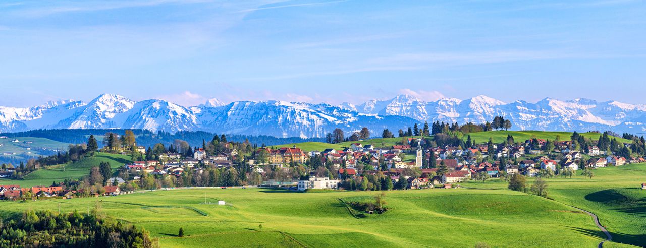 Bayerns schneebedeckte Berglandschaft im Hintergrund, davor grüne Hügel und kleine Dörfer