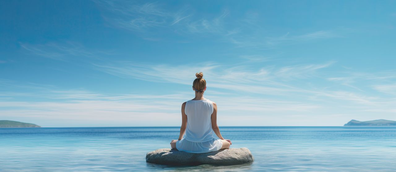 Eine Frau übt Ihre Yoga Asanas am Strand von Zypern. Sie sitzt auf einem Stein im Wasser und der Himmel strahlt schön blau.