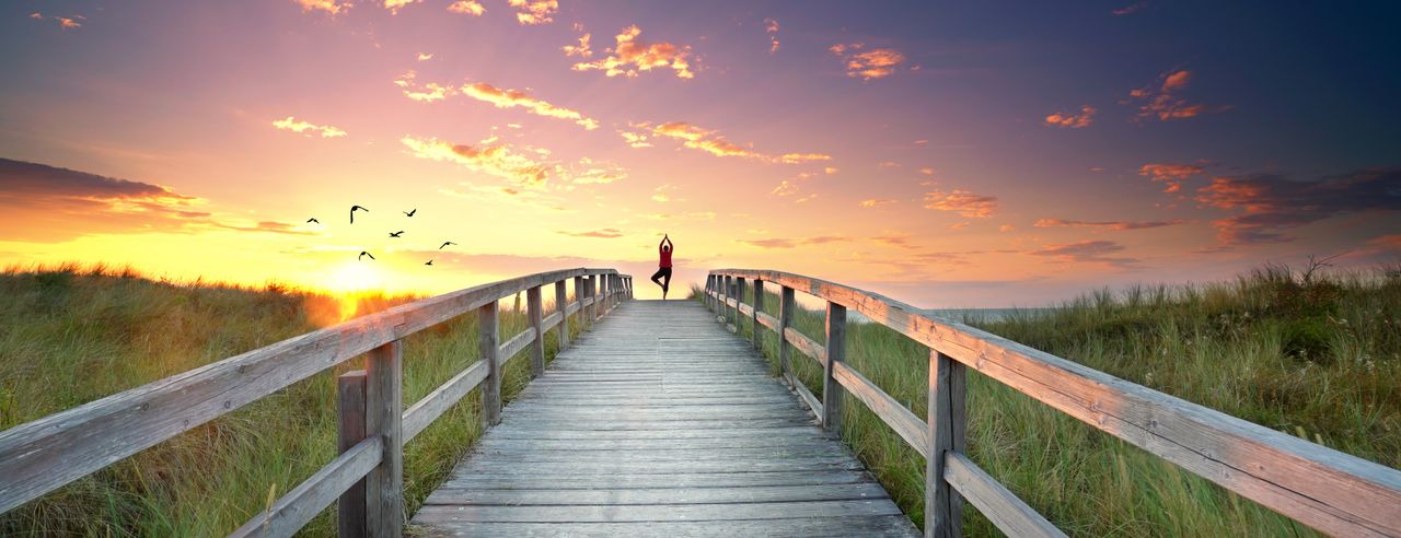 Eine Frau praktiziert Yoga auf dem Steg, der zum Strand führt. Die untergehende Sonne verfärbt den Himmel in glanzvolle Lichter.