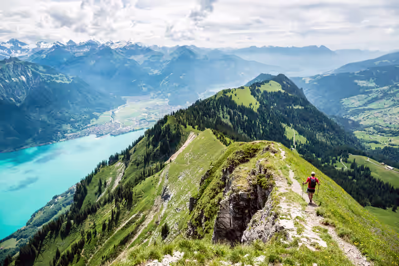 Wanderer mit Rucksack und Stöcken auf einem Bergkamm mit Aussicht auf ein Tal mit See, bewaldete Berge und schneebedeckte Alpengipfel