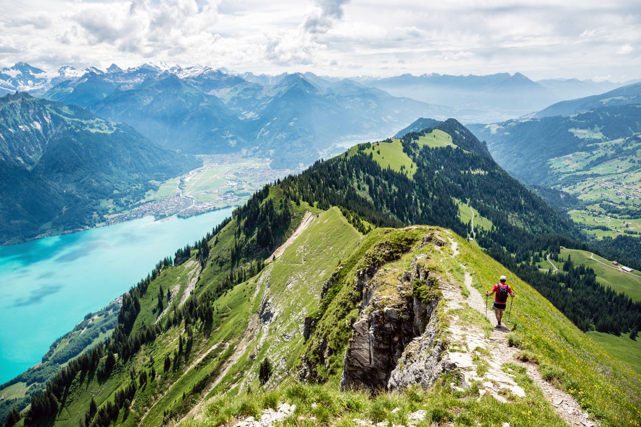 Wanderer mit Rucksack und Stöcken auf einem Bergkamm mit Aussicht auf ein Tal mit See, bewaldete Berge und schneebedeckte Alpengipfel
