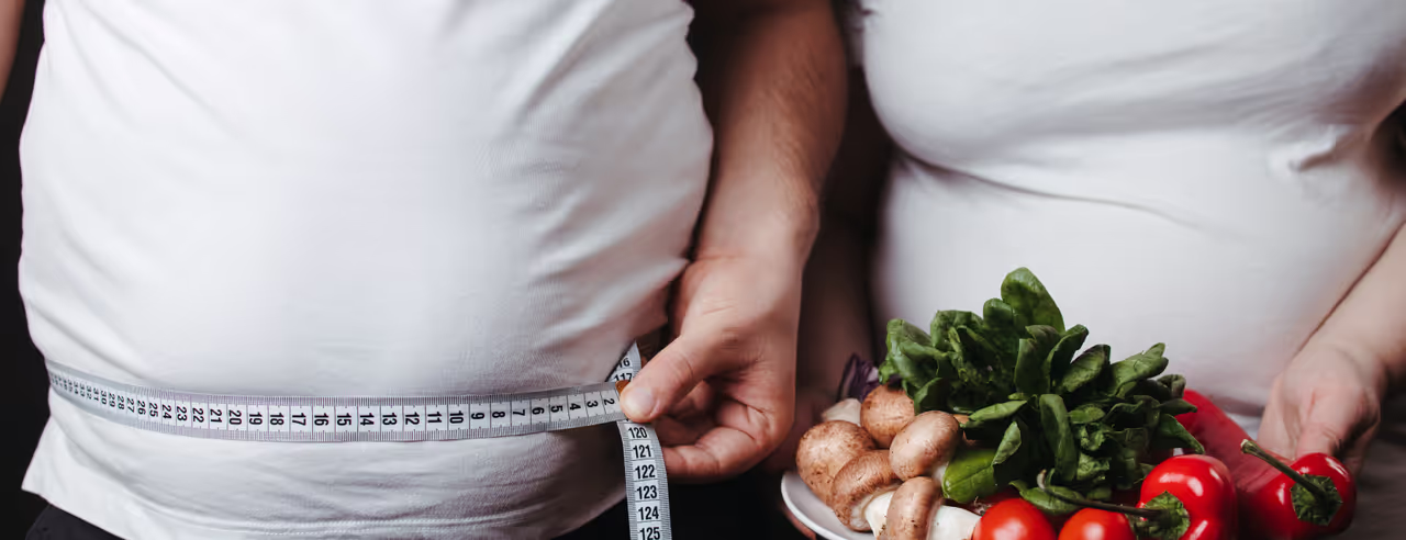 Paar mit Übergewicht in weißen T-Shirts und mit Maßband sowie Teller mit Gemüse darauf