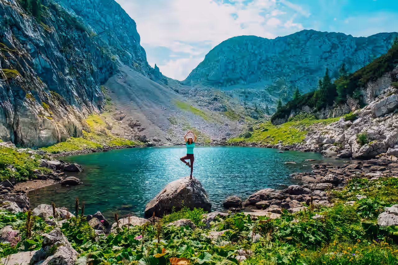 Eine Frau steht auf einem Felsen an einem türkisblauen See und macht eine Yoga-Pose
