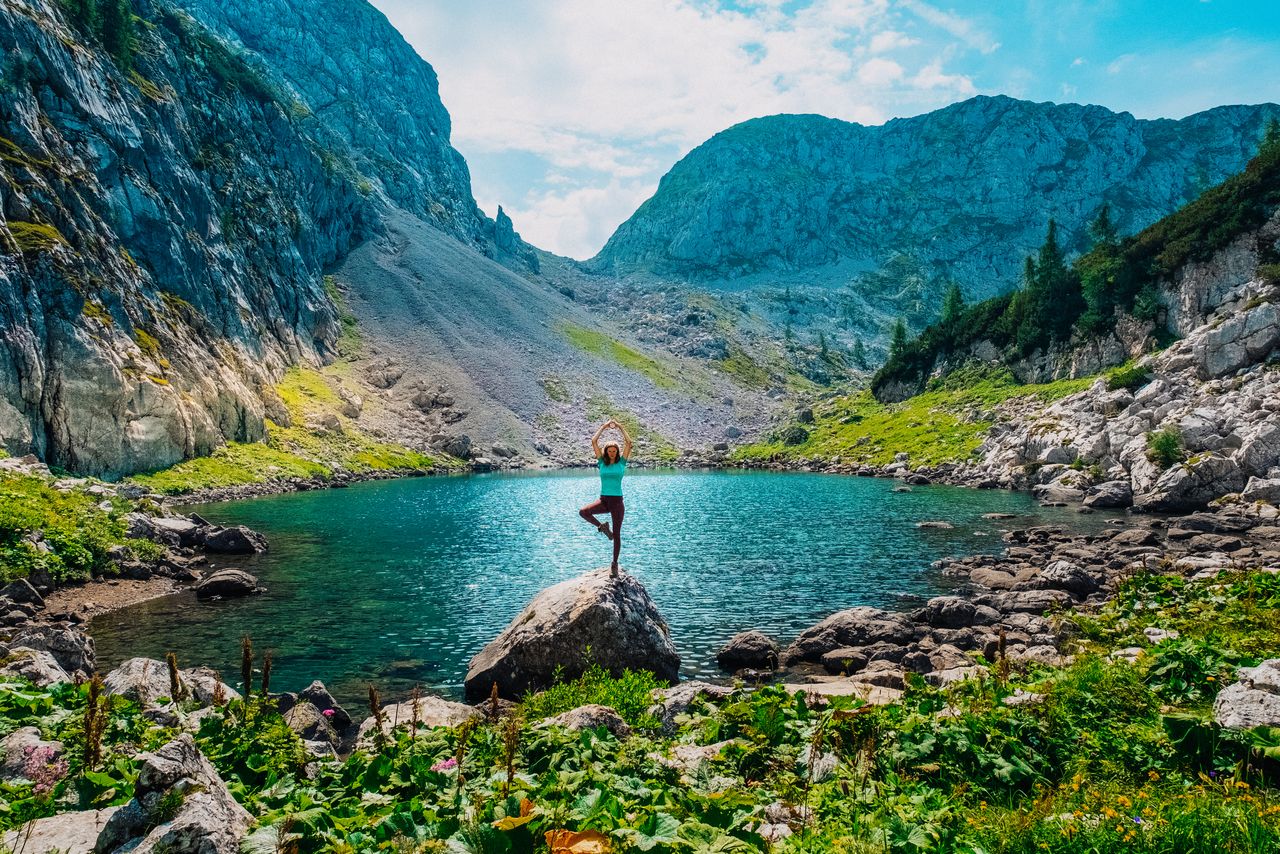 Eine Frau steht auf einem Felsen an einem türkisblauen See und macht eine Yoga-Pose