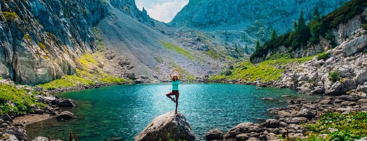 Eine Frau steht auf einem Felsen an einem türkisblauen See und macht eine Yoga-Pose