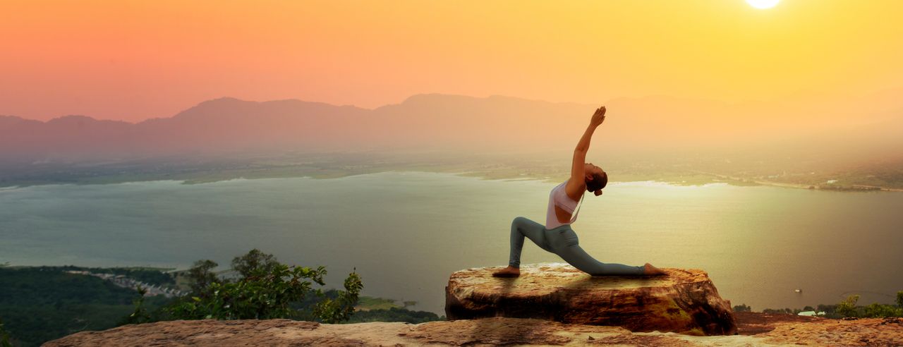 Frau auf einem Felsen in Yogastellung, im Hintergrund Meer und Sonnenuntergang
