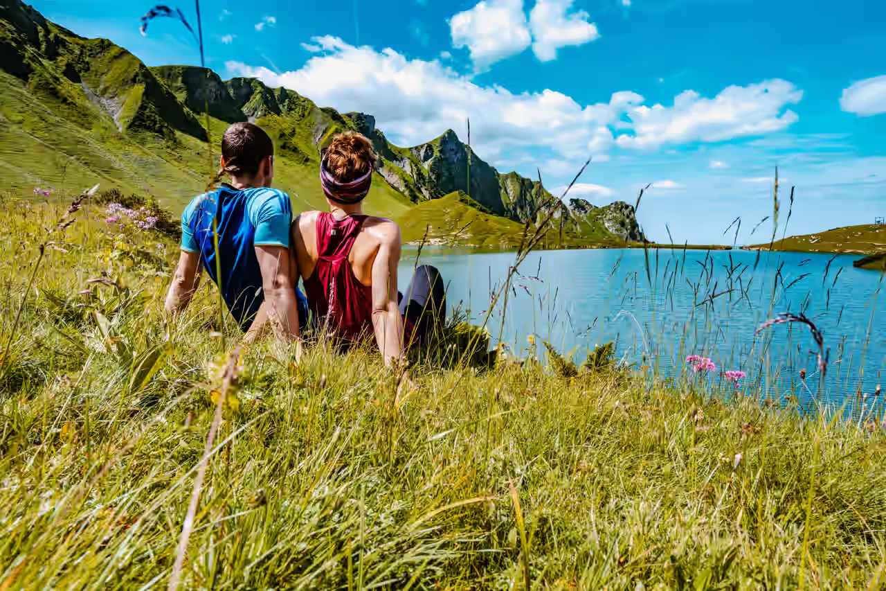 Ein Paar sitzt mit dem Rücken zu uns gedreht auf einer Blumenwiese und blickt auf die umliegende Natur.