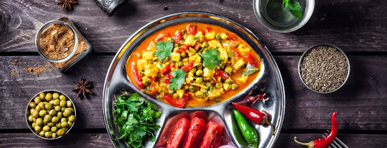 Auf einem schwarzen Tisch steht ein viergeteilter Teller, auf dem grünes, rotes, gelbes, lilafarbenes und orangefarbenes Essen zu sehen ist.