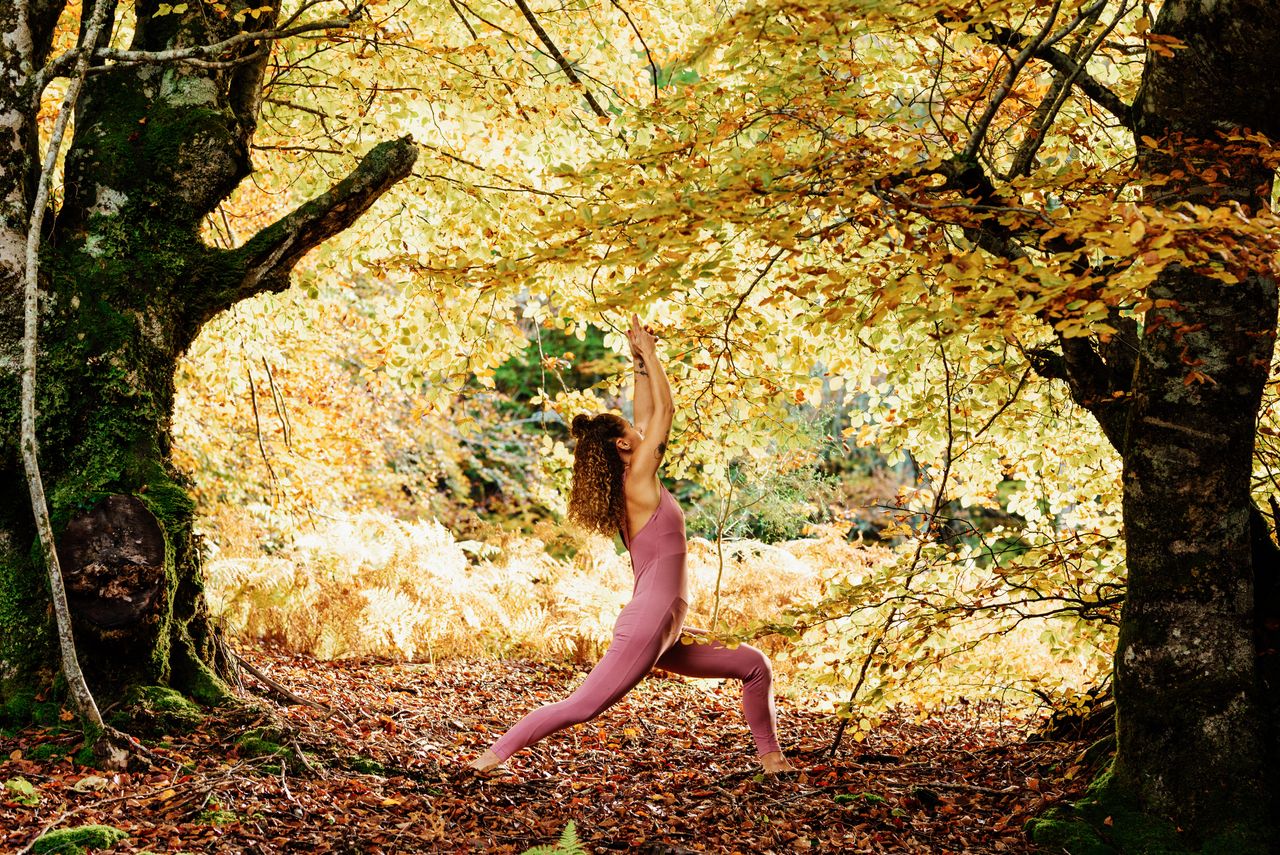 Eine Frau mit rosa Yogaanzug steht im Wald zwischen zwei Laubbäumen, deren Blätter herbstlich gelb leuchten und macht eine Yoga-Pose
