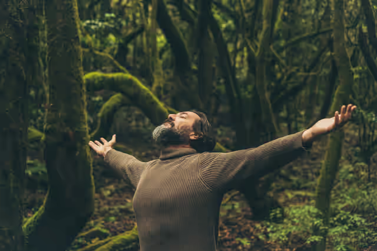 Ein Mann im braunen Pullover steht mit ausgestreckten Armen im Wald.