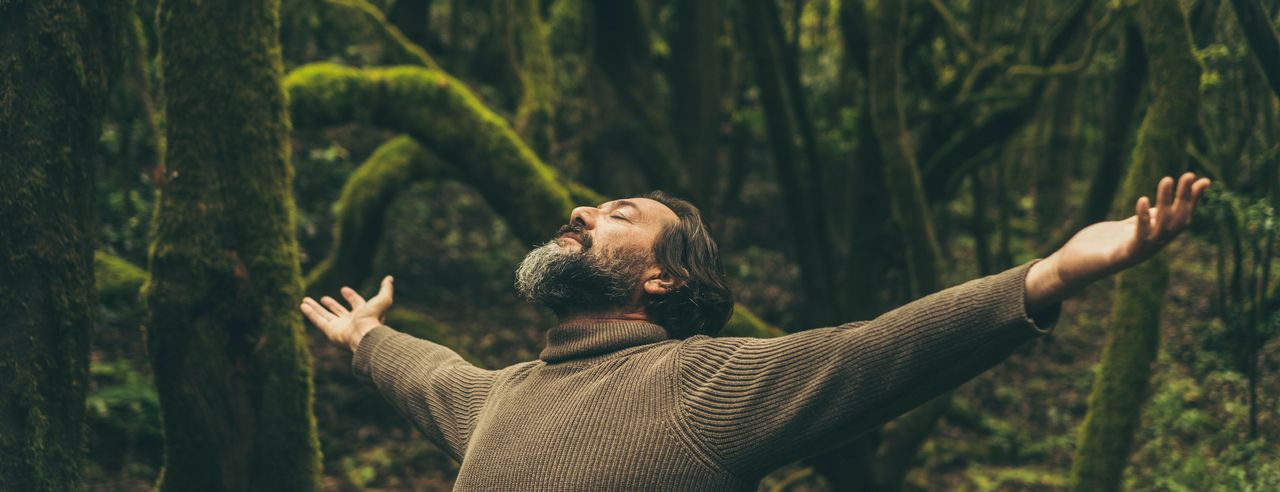 Ein Mann im braunen Pullover steht mit ausgestreckten Armen im Wald.