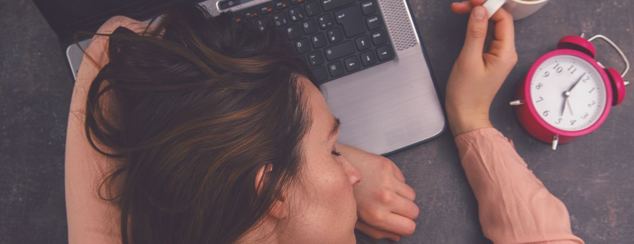 Eine Frau schläft auf einem Laptop. Dabei hält sie eine volle Kaffeetasse fest und eine pinker Wecker liegt neben ihr.