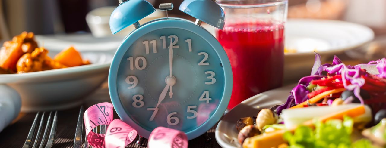 Blauer Wecker und rosafarbenes Maßband neben einem Teller mit Salat und einem Glas mit rotem Saft